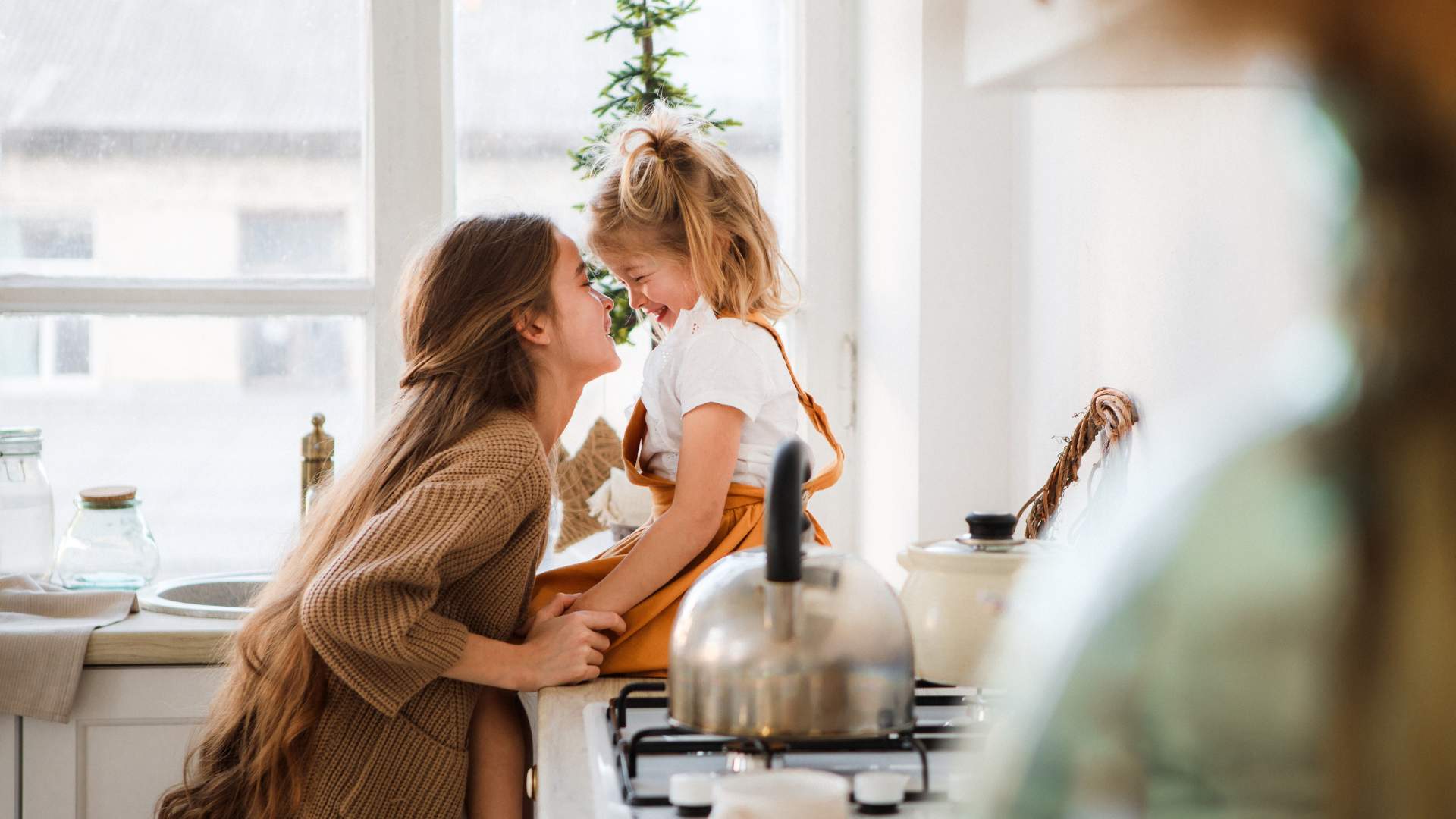 Two children in a kitchen smiling at each other, one sitting on the counter top and the other standing in front of them, with a teapot on the stove.
