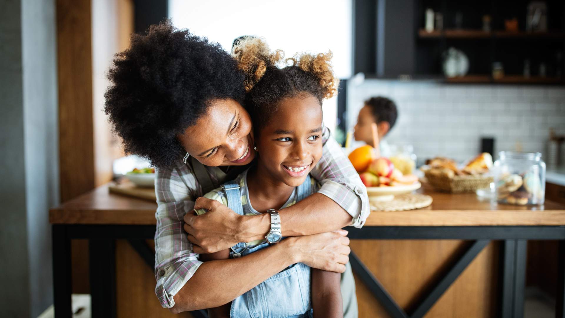 Woman hugging a young girl in a kitchen with fruit on the counter