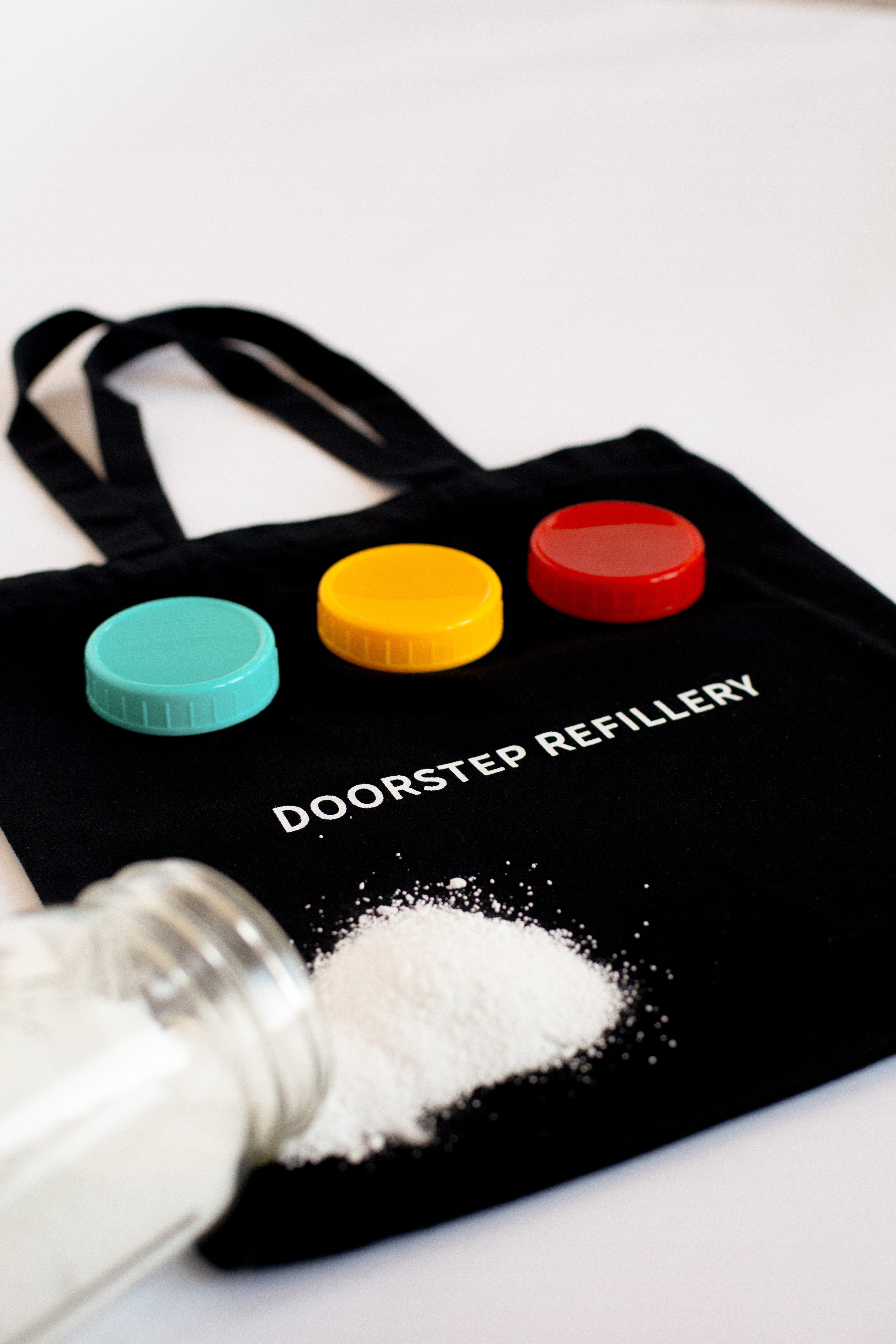 Black tote bag with colorful jar lids and 'Doorstep Refillery' text, with a white powdery cleaner spilling out.