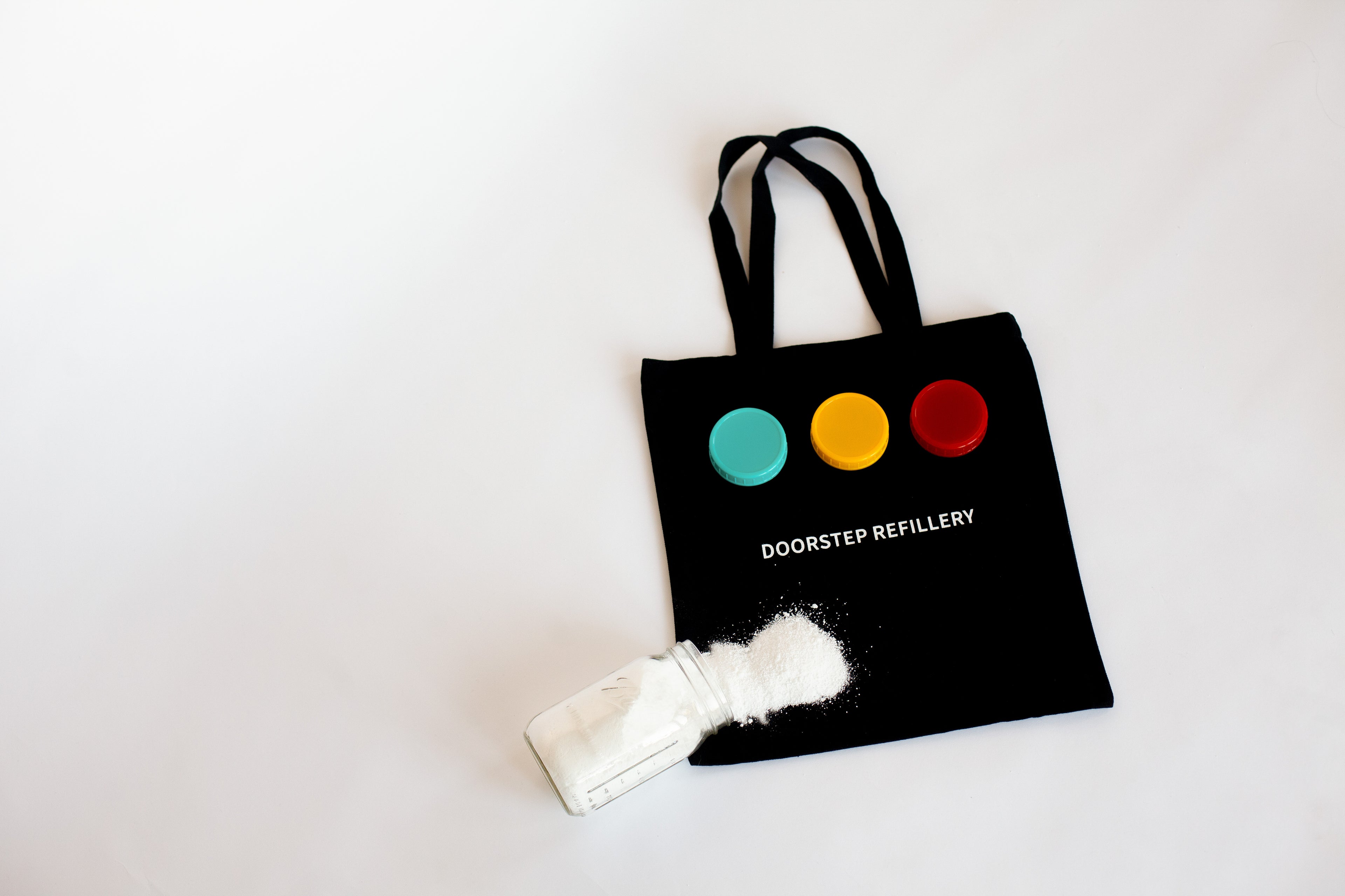 Black tote bag with colorful jar lids and 'Doorstep Refillery' text, accompanied by a jar of white powdered soap on a light gray background.