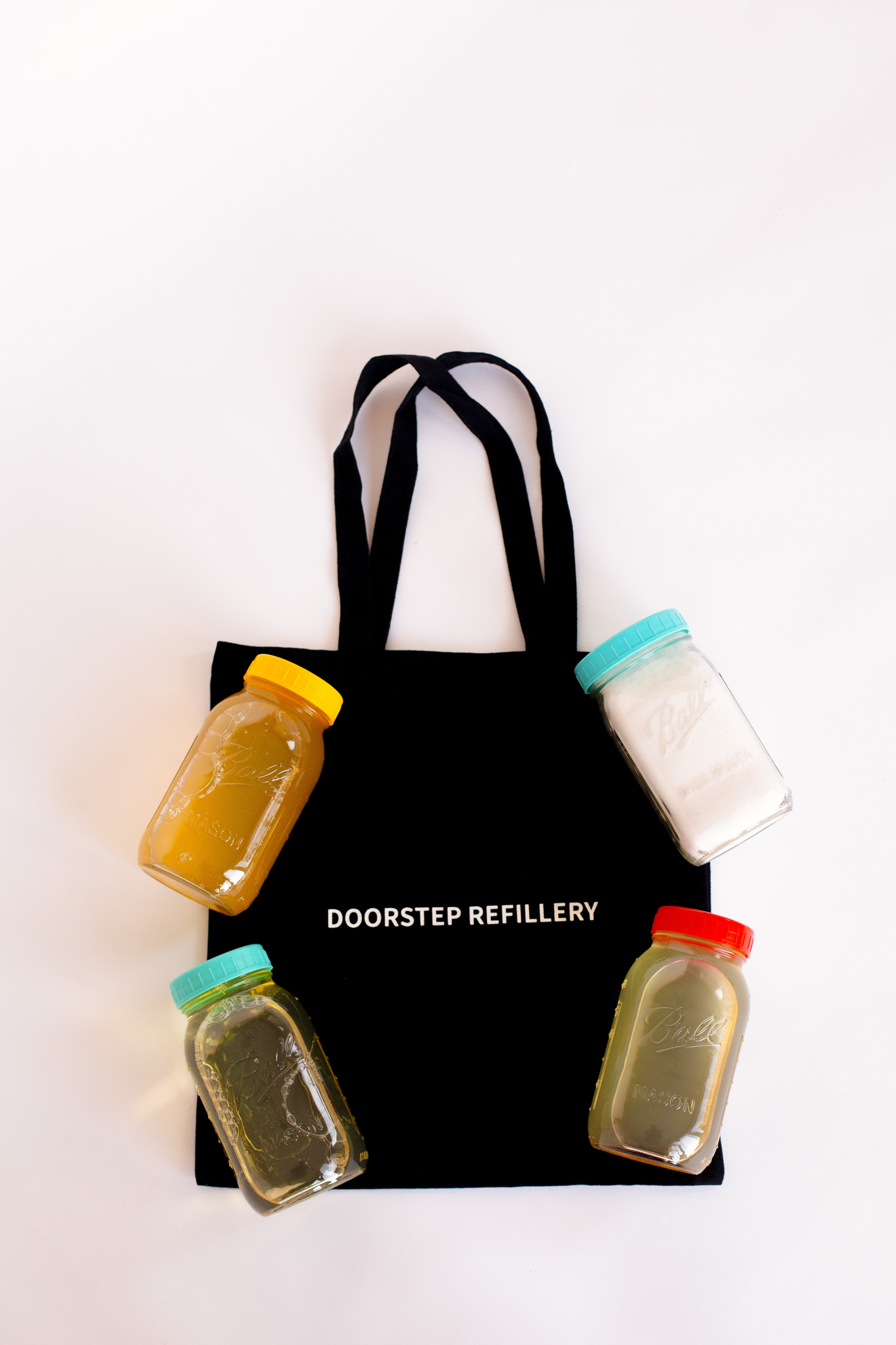 Black tote bag with 'Doorstep Refillery' branding and four refillable jars of various soaps on a white background